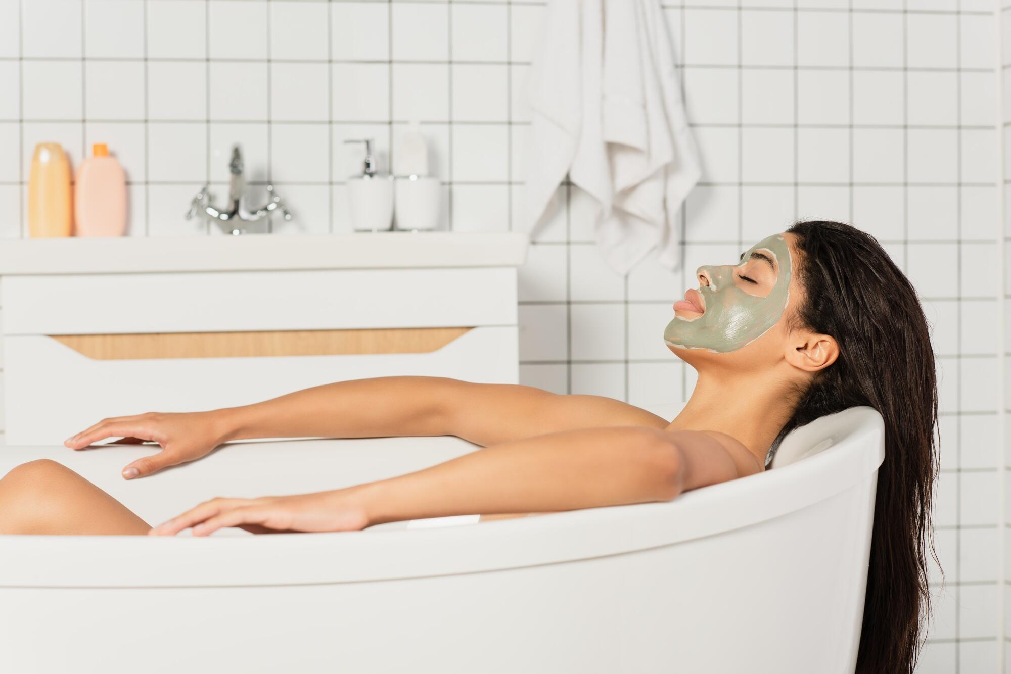 peaceful young african american woman with mud mask on face taking bath